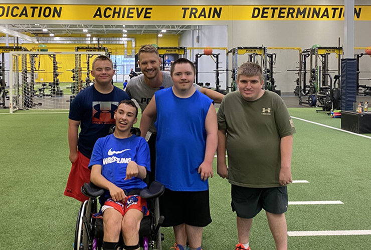 group of kids taking a picture in sanford sports bismarck facility. They are standing on turf and one of the kids is in a wheelchair with the coach standing behind all four of them