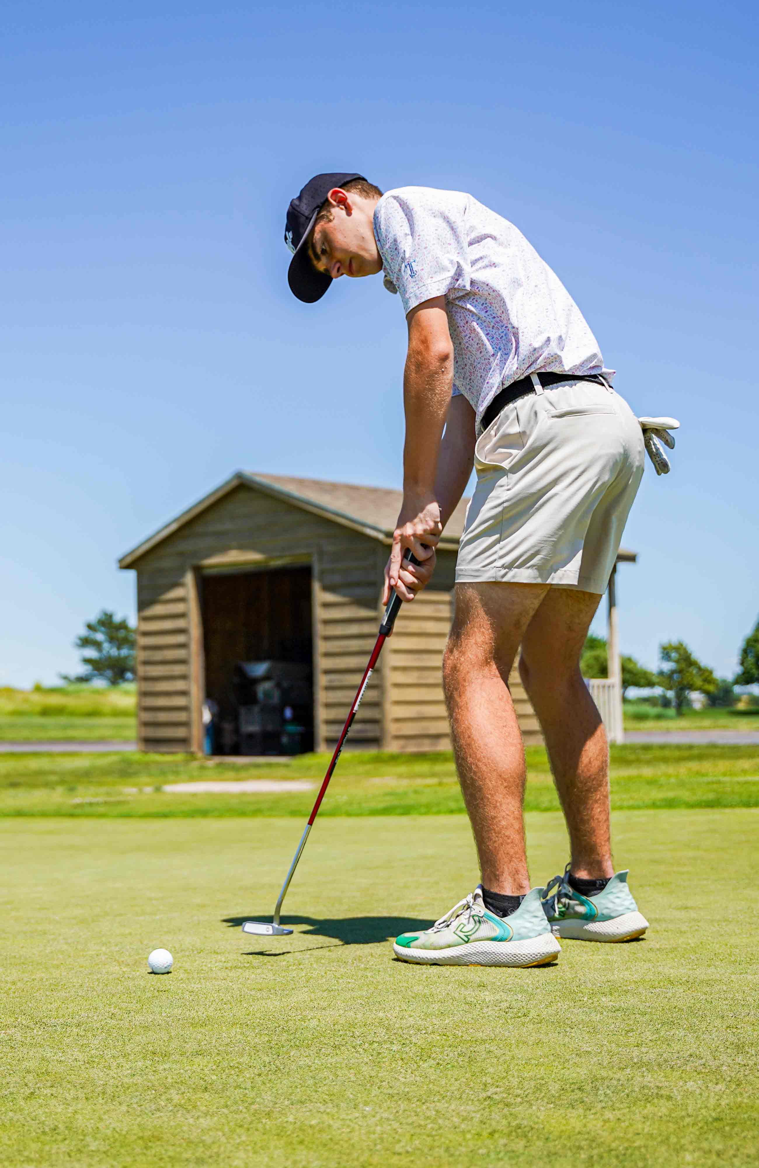 Golf player putting a golf ball, wearing golf attire with blue skies and a brown shed behind them