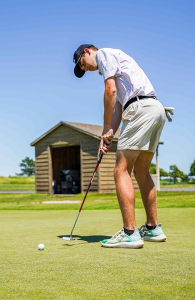 Golf player putting a golf ball, wearing golf attire with blue skies and a brown shed behind them