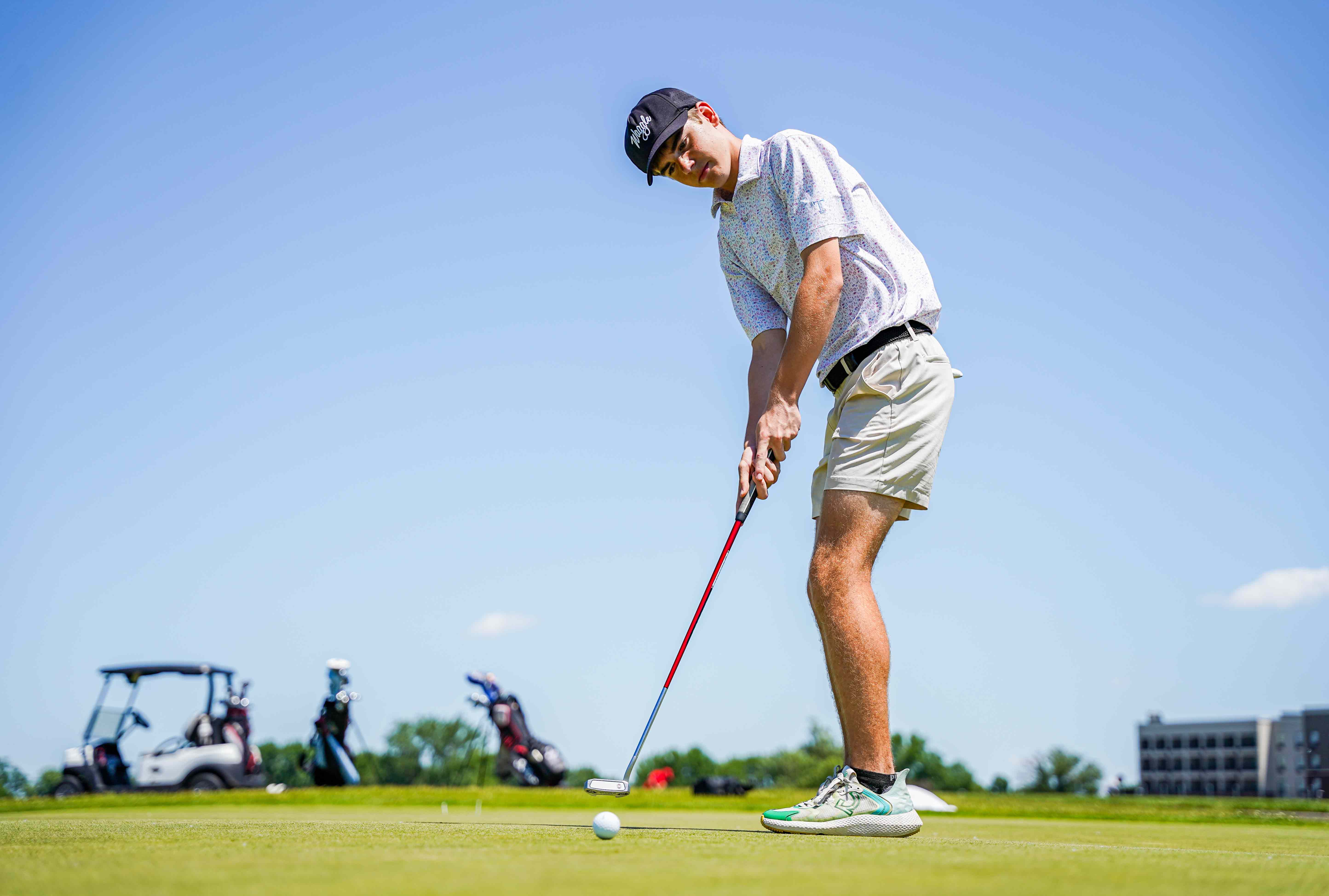 adult golf player outdoors hitting a golf ball in front of a blue sky and golf cart in the distance