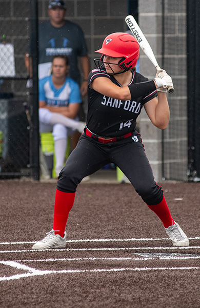 softball player wearing red and black jersey is winding up with a bat at home plate to hit ball